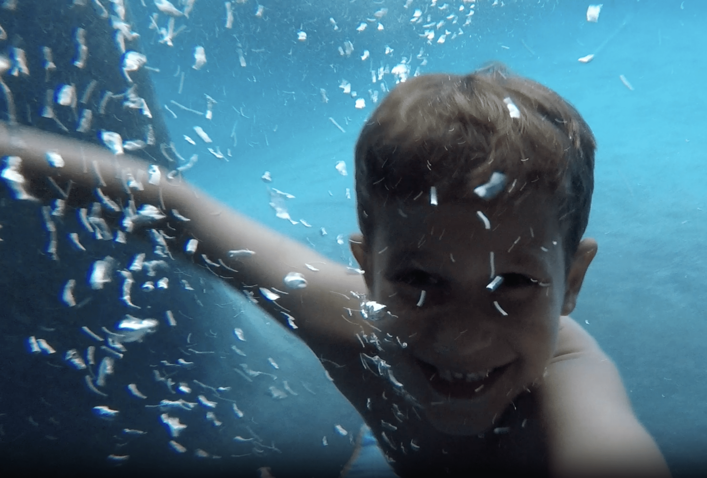 A joyful child is submerged in water, surrounded by bubbles, smiling at the camera in a playful, underwater photograph with a blue background.